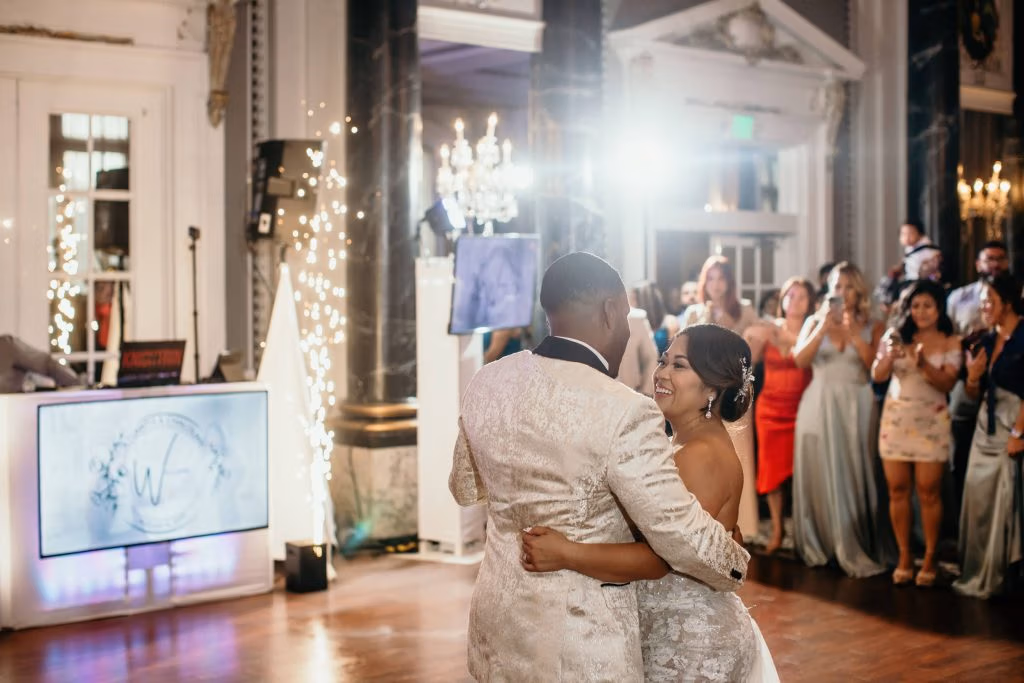 A bride and groom dance together on a ballroom floor while guests watch and take photos in an elegant, decorated venue.
