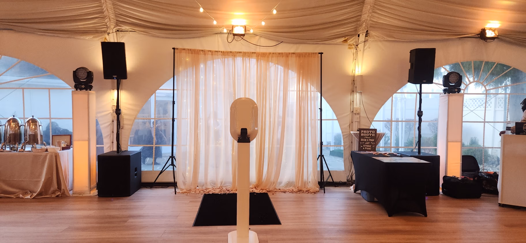 Photo booth setup inside a white tent with draped curtains, speakers, buffet tables on the left, and a black table with equipment on the right.