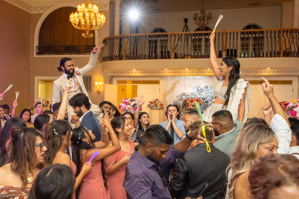 A large group of people dances and celebrates indoors, with two individuals raised above the crowd, holding glow sticks in a decorated event hall.